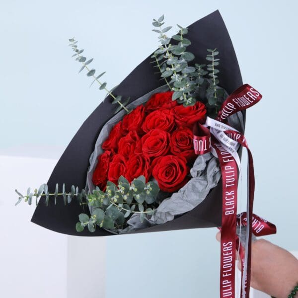 01 - Red Rose Romance Bouquet A hand-held bouquet of red roses with baby blue eucalyptus wrapped in black paper.