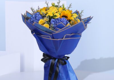 A bouquet of yellow roses, spray yellow roses, hydrangeas, delphiniums, eringium, alstroemeria, and oncidium wrapped in blue paper with a yellow ribbon.