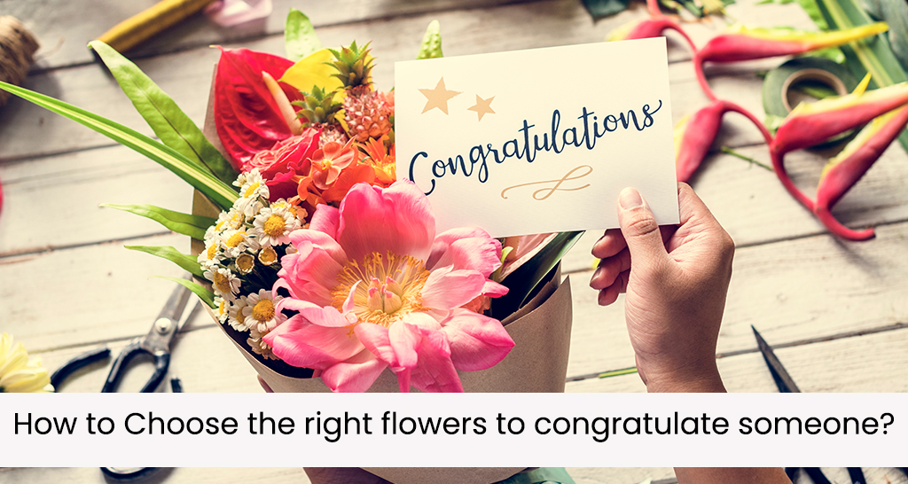 A person is holding a card with the word "Congratulations" written on it. The card is placed on top of a bouquet of colorful flowers, which includes pink peonies, white daisies, and other vibrant blooms. The flowers are arranged on a wooden table with various floral arrangement tools scattered around, such as scissors and tape. The text "How to Choose the right flowers to congratulate someone?" is displayed below the image.