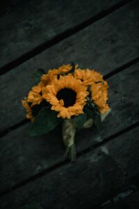 A bouquet of sunflowers sits on a dark wooden surface. The sunflowers are bright yellow with dark centers. There are also some smaller sunflowers and green leaves in the bouquet. The bouquet is tied with a yellow ribbon.