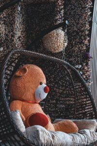 The image shows a large, brown teddy bear sitting on a wicker swing chair. The chair is hanging from chains and is surrounded by a dark, textured background.