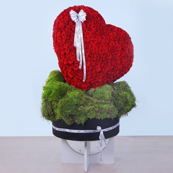 A large floral arrangement of 700 red roses in heart shape with a bow and greenery in round box tied with a white ribbon.