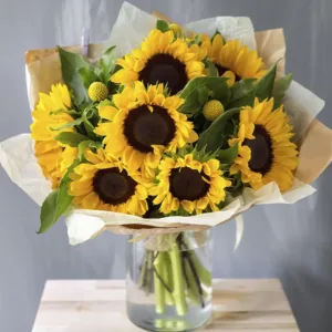 A bouquet of sunflowers is arranged in a clear glass vase, filled with water, and wrapped in light brown paper.