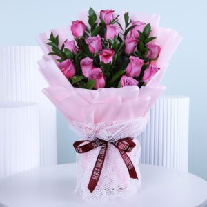 A beautifully arranged bouquet of purple roses wrapped in light pink paper and white lace, tied with a dark red ribbon.