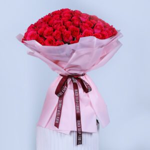 Bouquet of one hundred dark pink roses, wrapped in soft pink paper and secured with a dark maroon ribbon featuring decorative text.