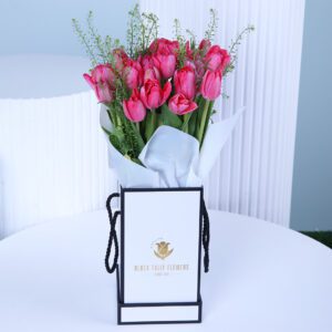 A vibrant bouquet of bright pink tulips arranged in a square white gift box with a black border and black rope handles.