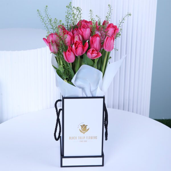 A vibrant bouquet of bright pink tulips arranged in a square white gift box with a black border and black rope handles.
