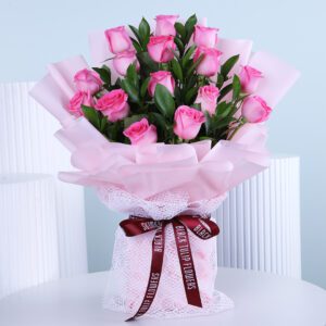 A bouquet of light pink roses wrapped in soft pink paper and delicate white netting, tied with a dark red ribbon.