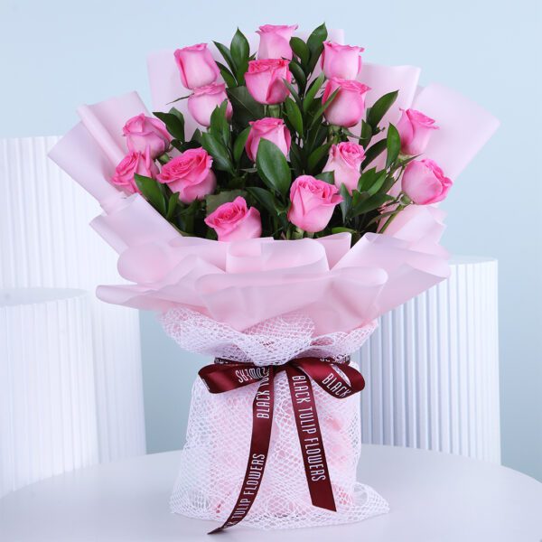 A bouquet of light pink roses wrapped in soft pink paper and delicate white netting, tied with a dark red ribbon.