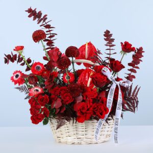 A white wicker basket filled with red roses, red anthuriums, red gerberas, eucalyptus, and textured foliage, finished with a Black Tulip Flowers branded ribbon