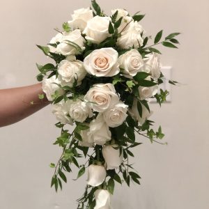 A cascading bridal bouquet A cascading bridal bouquet featuring numerous white and light pink roses, interspersed with lush green foliage, held by a hand.